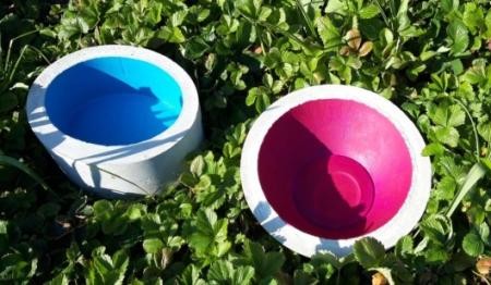  Zwei runde Betonschalen auf grünen Blättern, eine blau und eine pink bemalt.