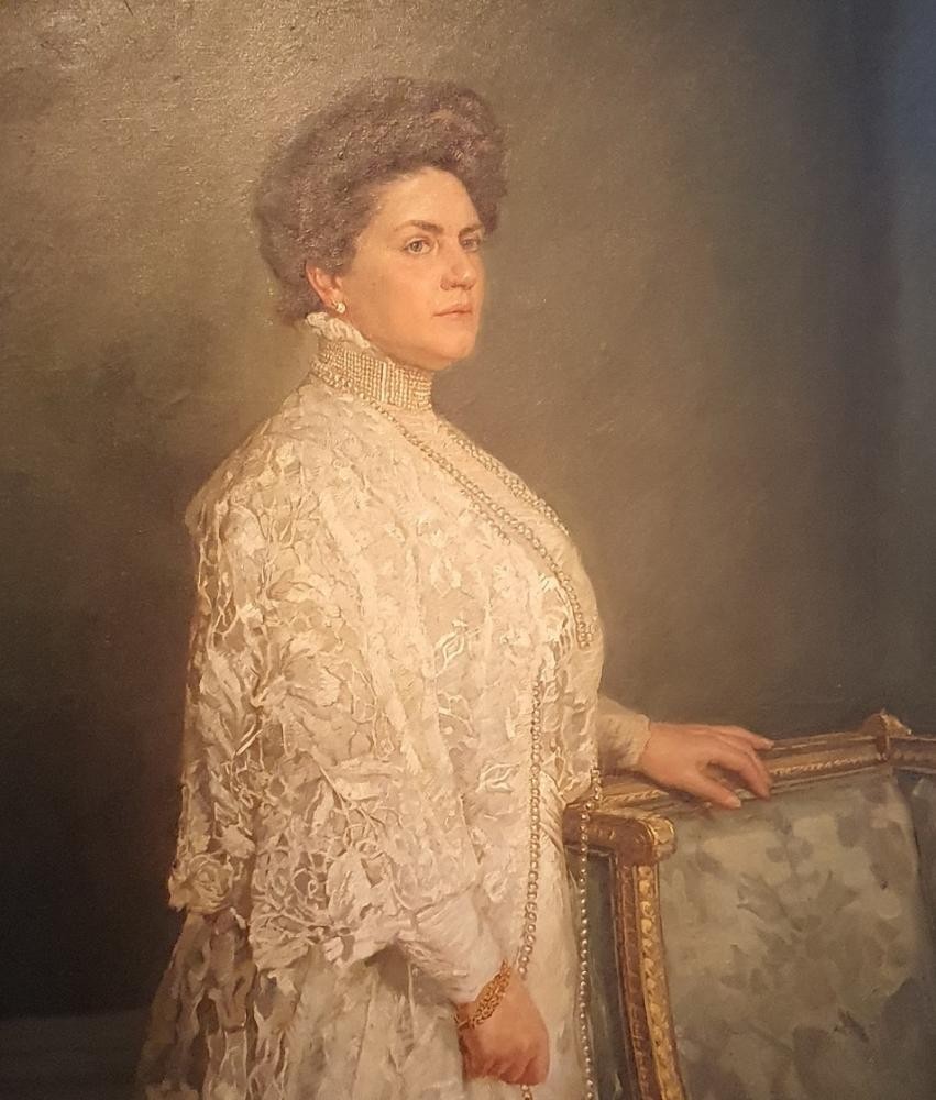 Charlotte von Württemberg.jpg Charlotte von Württemberg trägt ein weißes prächtiges Kleid und eine lange Perlenkette. Sie blickt in die Ferne und steht neben einem Sessel, ihre Hand ruht auf der Lehne.