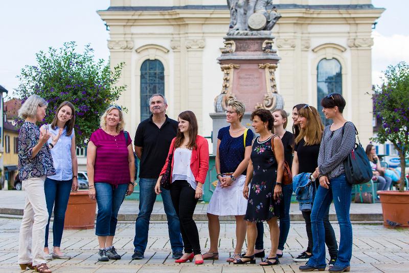 Eine Gruppe von Menschen steht vor dem Brunnen auf dem Marktplatz und schaut und hört gespannt einer Gästeführerin zu.