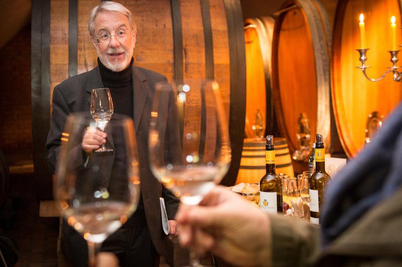 Im Vordergrund sieht man zwei Hände, die Weingläser halten, dahinter ein Mann im Sakko und Brille, der auch ein Glas hält. Im Hintergrund stapeln sich Weinfässer.