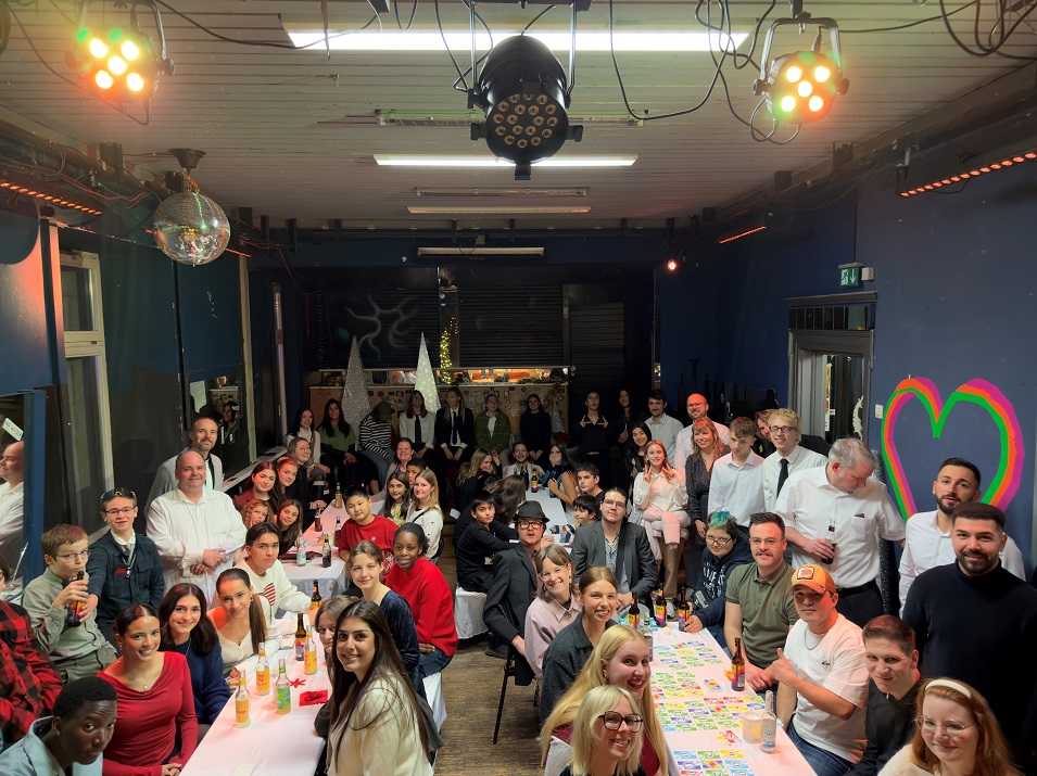 Etwa 60 Personen (Jugendliche und Erwachsene) sitzen an langen Tischen bzw. stehen dahinter und lächeln in die Kamera.