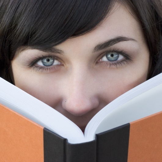 Frau mit schwarzen Haaren und blauen Augen schaut hinter einem Buch hervor