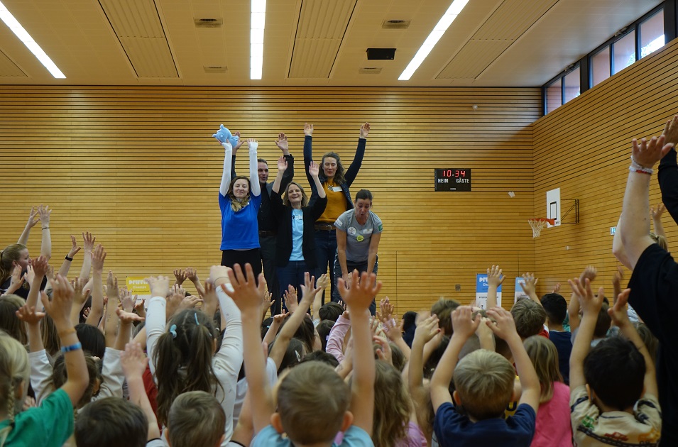 In einer großen Sporthalle stehen fünf Personen, ein Mann und vier Frauen, auf einer kleinen Bühne und heben fröhlich die Arme in die Luft. Vor ihnen steht eine große Gruppe von Kindern, die ebenfalls die Arme hochstreckt und begeistert mitmacht.