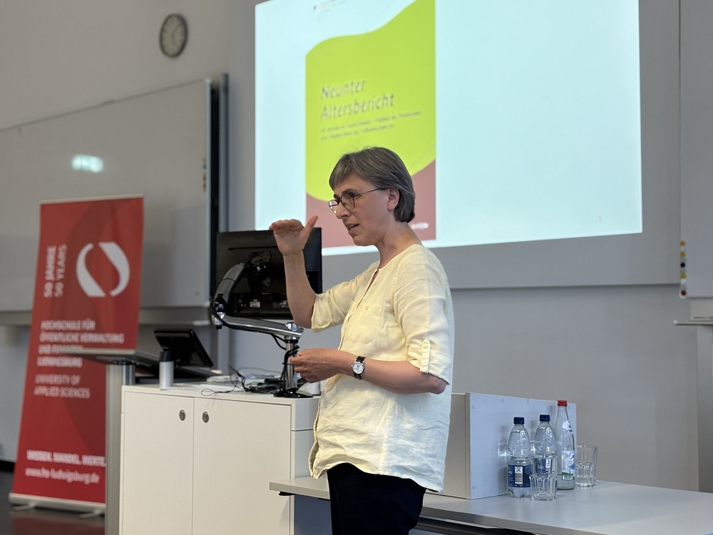 Frau Gaugel hält einen Vortrag zum „Neusten Altersbericht“ vor Präsentationsfolie, mit Jubiläumsbanner der Hochschule für öffentliche Verwaltung und Finanzen Ludwigsburg im Hintergrund.