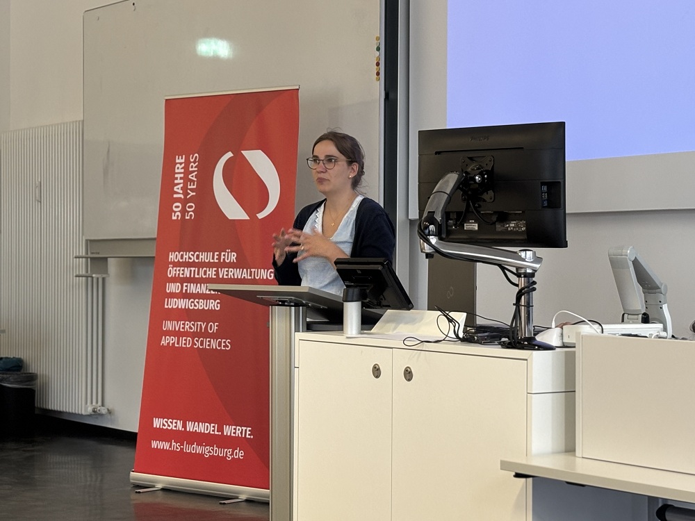 Frau Clauss hält einen Vortrag zum "Neuesten Altersbericht", mit Jubiläumsbanner der Hochschule für öffentliche Verwaltung und Finanzen Ludwigsburg im Hintergrund