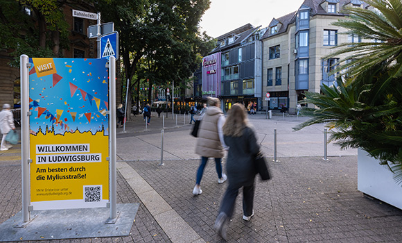 Auf einer Stele steht: "Willkommen in Ludwigsburg. Am besten durch die Myliusstraße!" Im Hintergrund befindet sich die Myliusstraße.