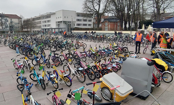 Fahrräder in mehreren Reihen. Im Vordergrund bunte Kinderräder. An einem Stand stehen mehrere Menschen in orangenen Warnwesten.