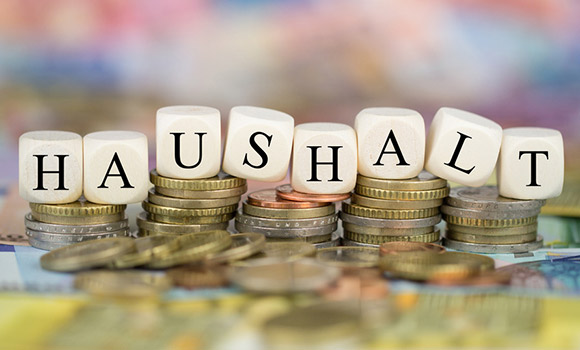 Auf Stapeln mit Münzen liegen acht Würfel. Auf jedem Würfel steht ein Buchstabe. Diese bilden das Wort "Haushalt".