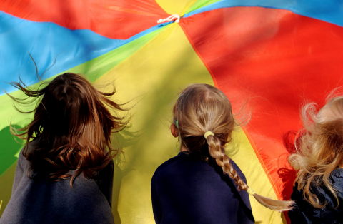 Drei Kinder schauen auf einen bunten Fallschirm mit Abschnitten in Rot, Blau, Grün und Gelb.