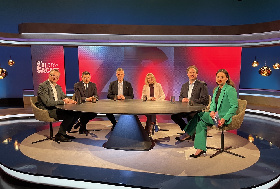 sechs Personen sitzen in einem Fernsehstudio um einen Tisch. 
