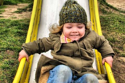 Ein Kind mit grüner Mütze rutscht eine gelbe Rutsche auf einem Spielplatz hinunter.