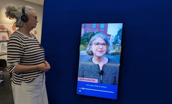Frau mit Kopfhörern schaut auf einer Leinwand einen Film. In diesem spricht gerade eine Frau.