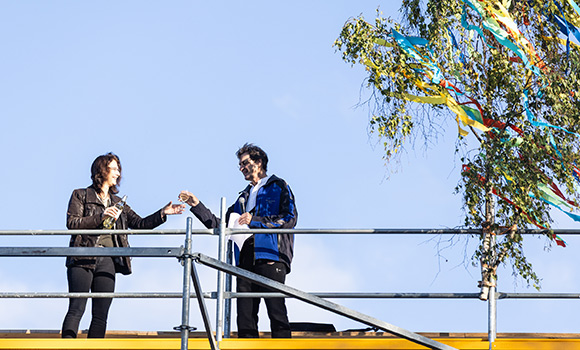Baubürgermeisterin Andrea Schwarz und Zimmermann Frank Grottenmüller stehen auf dem Dach der Oststadthalle und stoßen an. Neben ihnen hängt die Richtkrone.