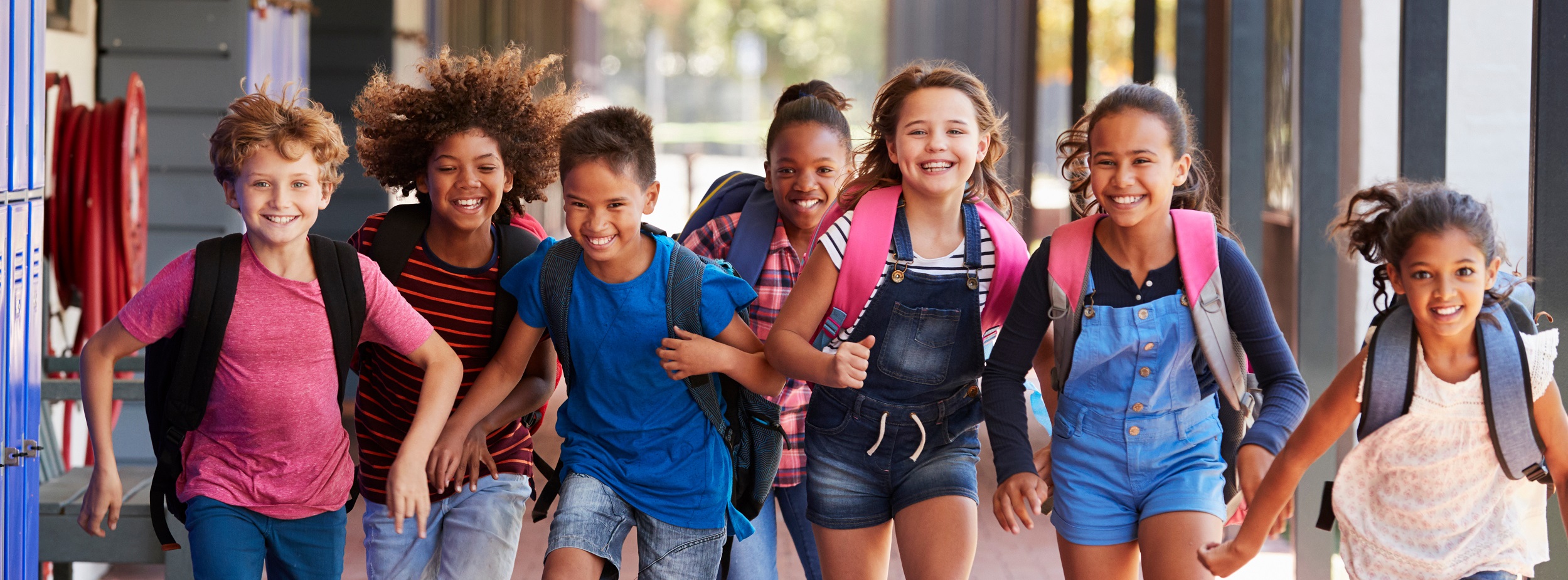 Sieben Kinder mit Rucksäcken laufen zusammen in einem Außenkorridor.
