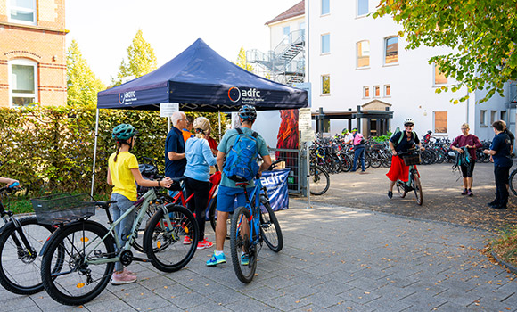 Mehrere Fahrradfahrer stehen an einem Pavillon des ADFC. Hinter dem Pavillon befindet sich ein Hof, auf dem viele Fahrräder geparkt sind.