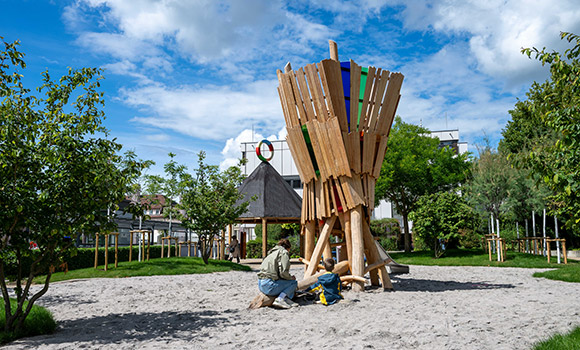 Kinder spielen mit ihrer Mutter im Sand vor einem Spielturm aus Holz. Im Hintergrund steht eine Hütte. Bäume und Rasenhügel befinden sich ebenfalls auf der Fläche.
