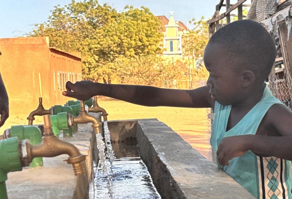 Ein Kind dreht an einem Wasserhahn eines Trinkwasserbrunnens, während klares Wasser in ein Becken fließt.