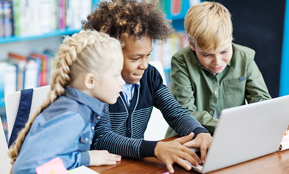 Ein Mädchen und zwei Jungen sitzen gemeinsam vor einem Notebook. Der Junge in der Mitte schreibt etwas; die anderen Kinder schauen auf den Bildschirm.