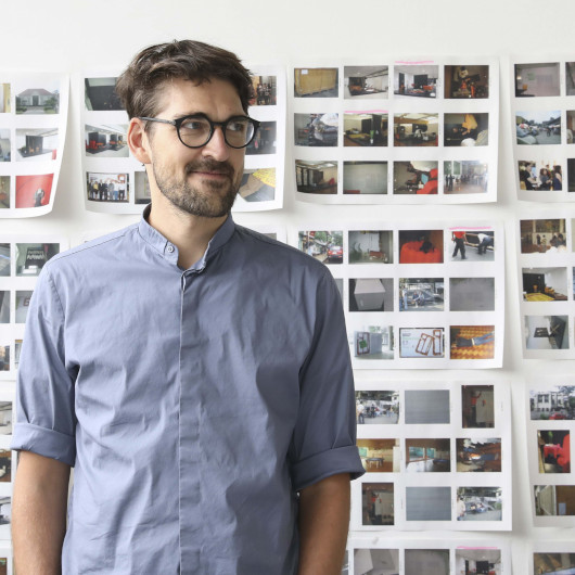 Ein Mann mit blauem Hemd und schwarzer Brille steht vor einer Wand mit vielen kleinen Fotografien.