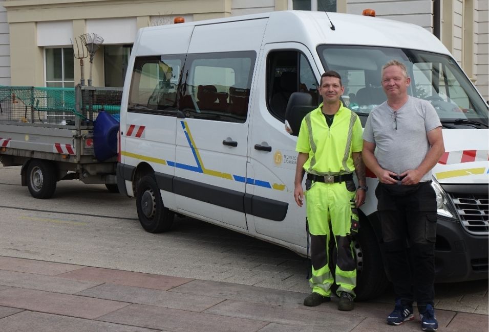 Zwei Männer stehen vor einem Bus mit Anhänger. Der Mann links trägt gelbe Arbeitskleidung.