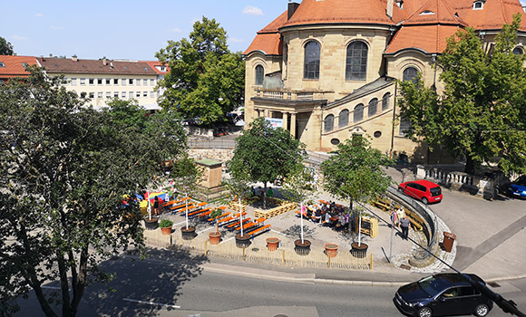 Vor der Friedenskirche ist eine ovale Fläche mit Bäumen und Sitzmöbeln angelegt worden. Vor dem kleinen Platz fährt auf einer Straße ein Auto vorbei.