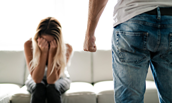 Eine Frau sitzt auf dem Sofa, die Hände hält sie vor dem Gesicht. Ein Mann steht vor dem Sofa, seine Hand ist zur Faust geballt.