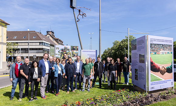 Rund 25 Menschen stehen auf der Mittelinsel einer Straßenkreuzung. Sie ist mit Rasen und Blumen bepflanzt. Außerdem stehen auf der Kreuzung Plakatwände mit Informationen.