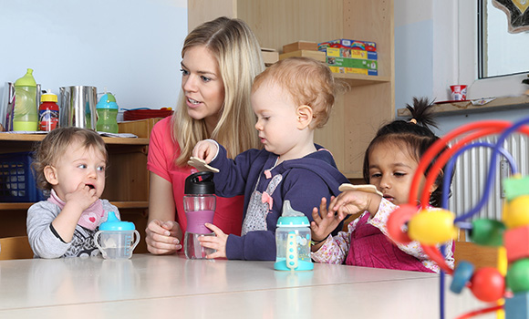 Drei Kleinkinder und eine Frau sitzen an einem Tisch. Auf dem Tisch stehen Trinkflaschen.