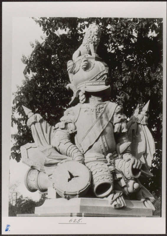 Eine Person kniet auf einem Rollgerüst vor einer großen weißen Stoffbahn mit aufgenähten Taschen. Davor steht eine Person und liest.