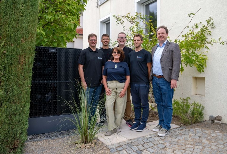 Sechs Menschen stehen vor einem Gebäude, links von ihnen die Wärmepumpe