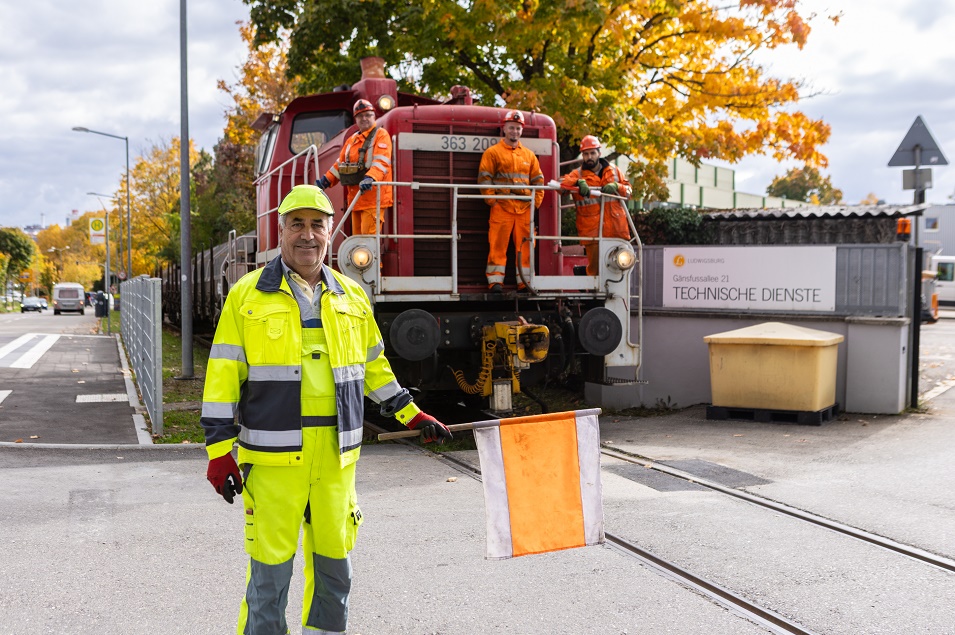 Ein Mann in gelber Warnkleidung und mit einer Fahne in der Hand steht vor einem Zug. Auf dem Zug stehen drei weitere Männer in orangener Kleidung.