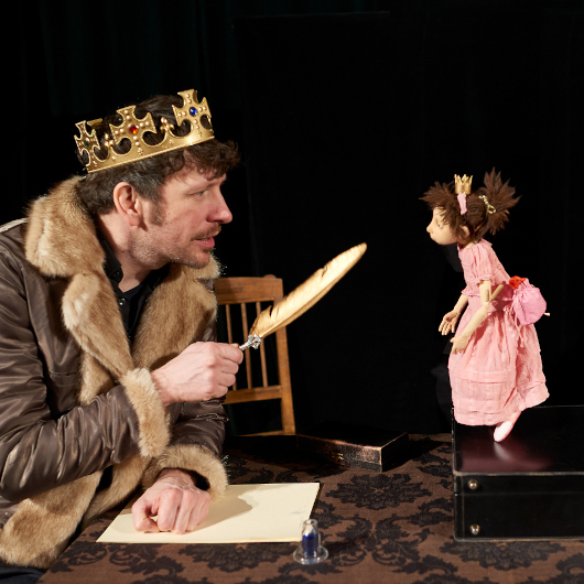 Ein als König verkleideter Mann sitzt einer Prinzessinenpuppe auf einer Bühne gegenüber. 