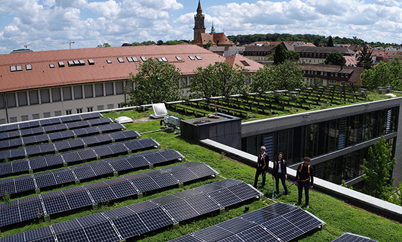 Auf dem begrünten Dach befinden sich eine Vielzahl an Solarmodulen. Drei Personen stehen angegurtet auf dem Dach.