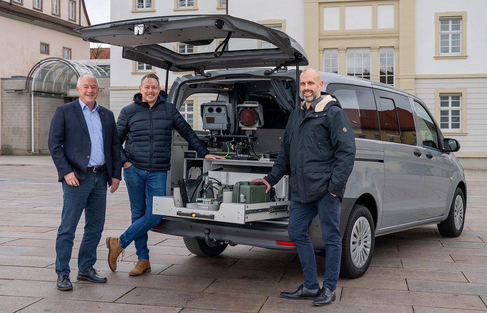 Drei Männer stehen an einem Auto mit offenem Kofferraum. Darin sind technische Geräte zu sehen.
