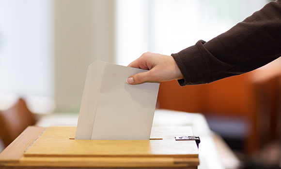 Eine Person, von der man nur den Arm sieht, steckt einen Stimmzettel in eine Wahlurne.