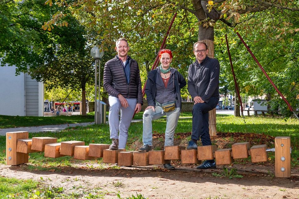 Drei Menschen stehen in einem Park hinter einem Spielgerät mit Klötzen aus Holz.