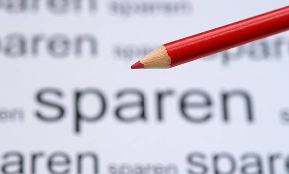Roter Buntstift vor einer weißen Fläche, auf der mehrfach das Wort "sparen" steht