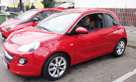 Zwei rote Carsharing-Autos stehen nebeneinander in zwei Parkboxen. In einem der Autos sitzt eine Frau auf dem Fahrersitz.