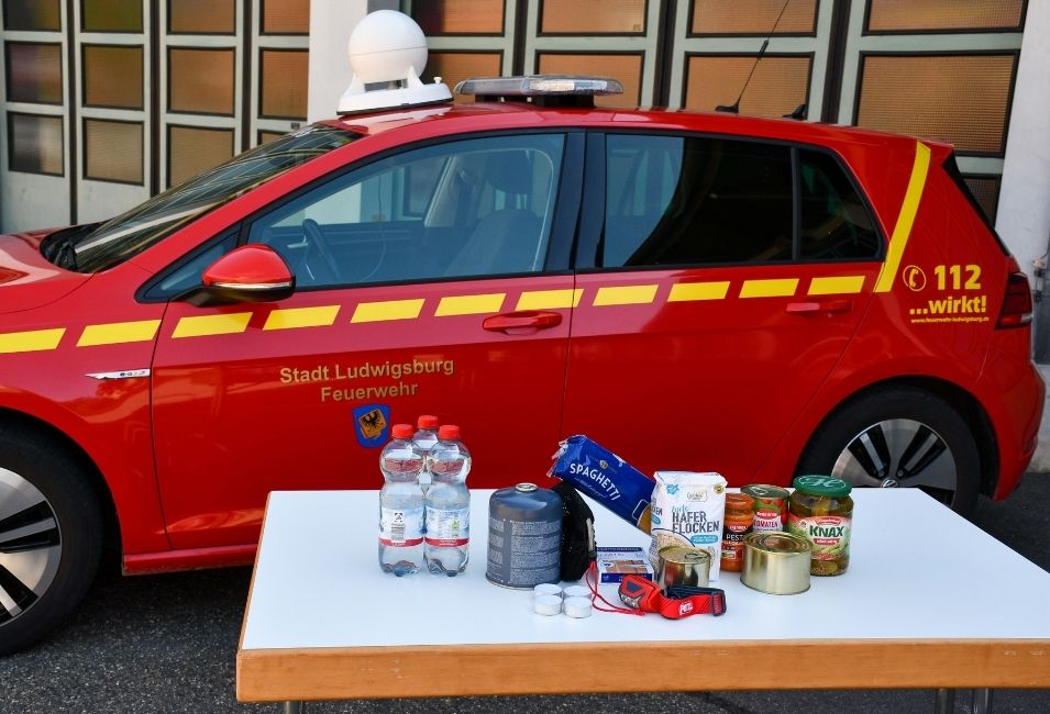 Vor einem roten Einsatzfahrzeug der Feuerwehr Ludwigsburg stehen auf einem Tisch verschiedene Gegenstände für die Notfallvorsorge. Zu sehen sind unter anderem Wasserflaschen, haltbare Lebensmittel, ein Campingkocher, Teelichter und eine Stirnlampe.