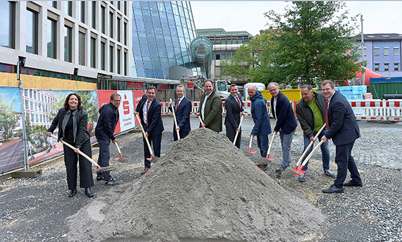 Die zehn Personen stehen auf dem Vorplatz der Kreissparkasse im Halbkreis. Vor ihnen befindet sich ein kleiner Sandhaufen, auf den sie mit je einem Spaten mehr Sand schaufeln.