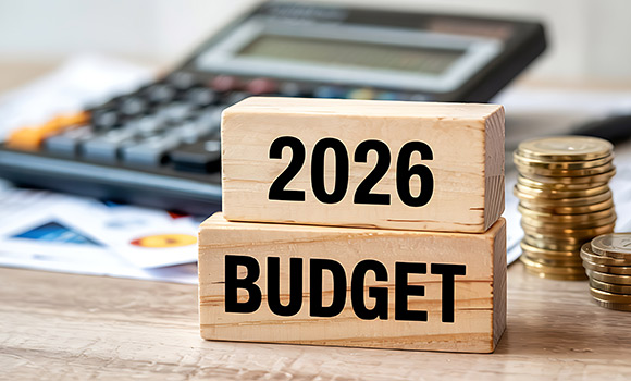 Holzklötze mit der Aufschrift "2026 Budget". Dahinter Taschenrechner und ein Stapel mit Münzen.