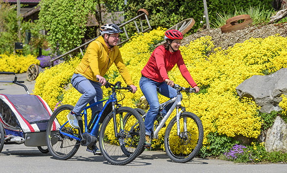 Ein Mann und eine Frau fahren zusammen mit dem Rad. Am Fahrrad des Mannes befindet sich ein Fahrradanhänger, in dem ein Kleinkind sitzt.