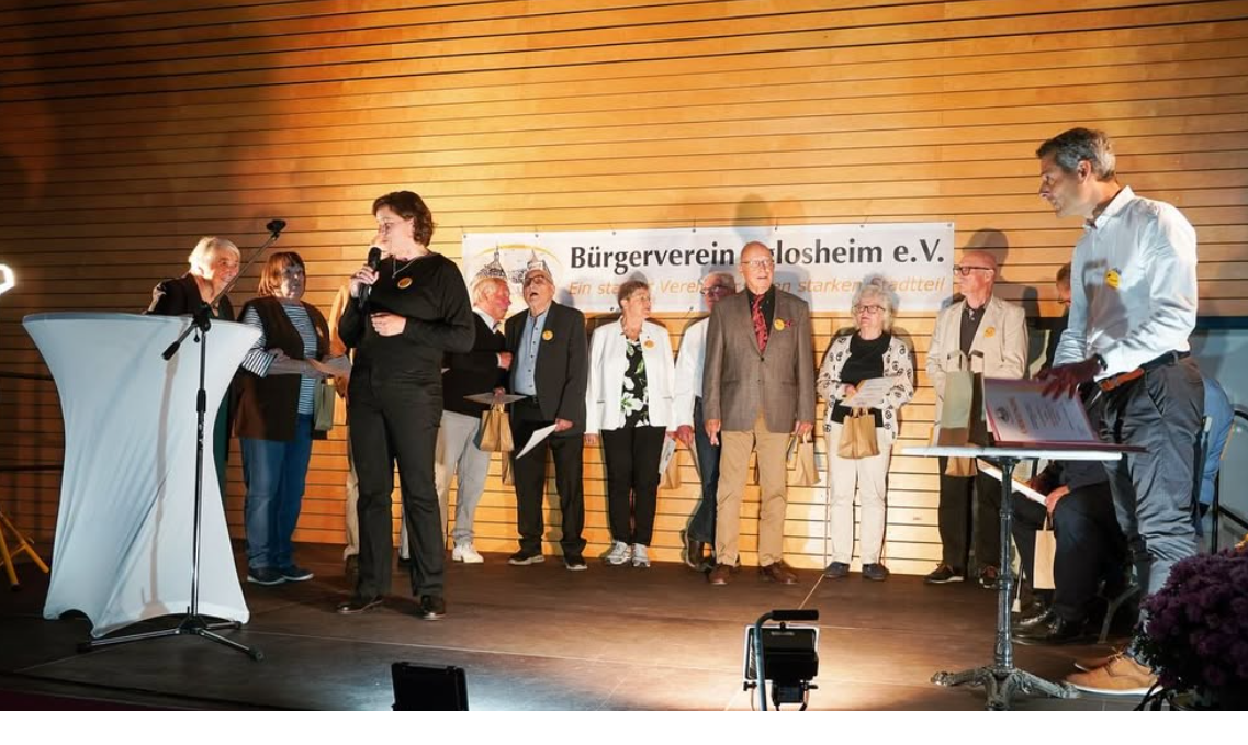 Personen auf einer Bühne. Eine Frau spricht im Mikrofon.