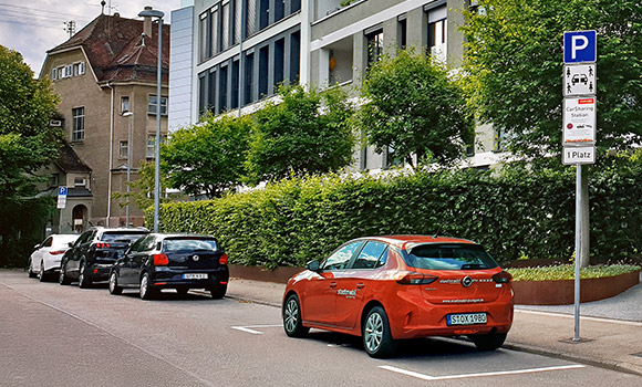 Ein rotes stadtmobil-Fahrzeug steht auf einem Parkplatz an einer Straße. Davor stehen mehrere private Pkw.