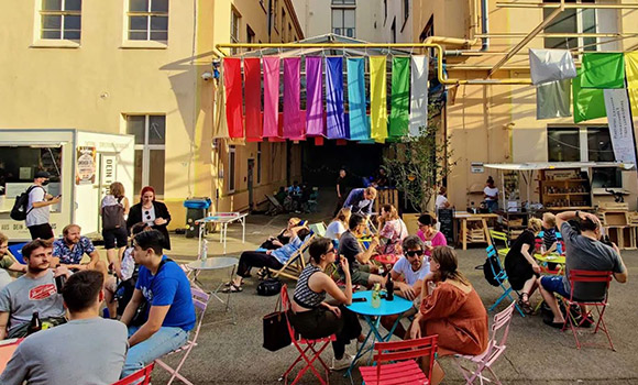 Menschen sitzen an Tischen. Im Hintergrund steht ein Food-Truck.