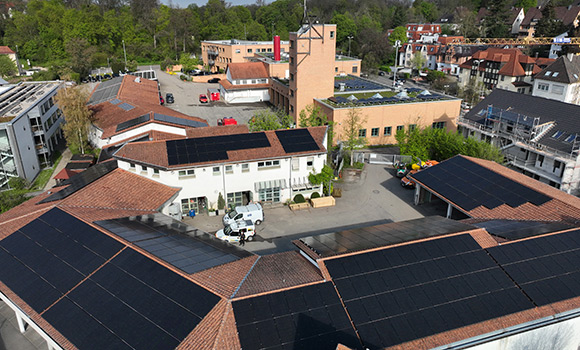 Luftaufnahme der Photovoltaikanlagen auf dem Flachdach der Hauptfeuerwache, dem Satteldach des benachbarten TDL-Gebäudes und einer Containerbedachung auf dem Hof der Feuerwehr.  