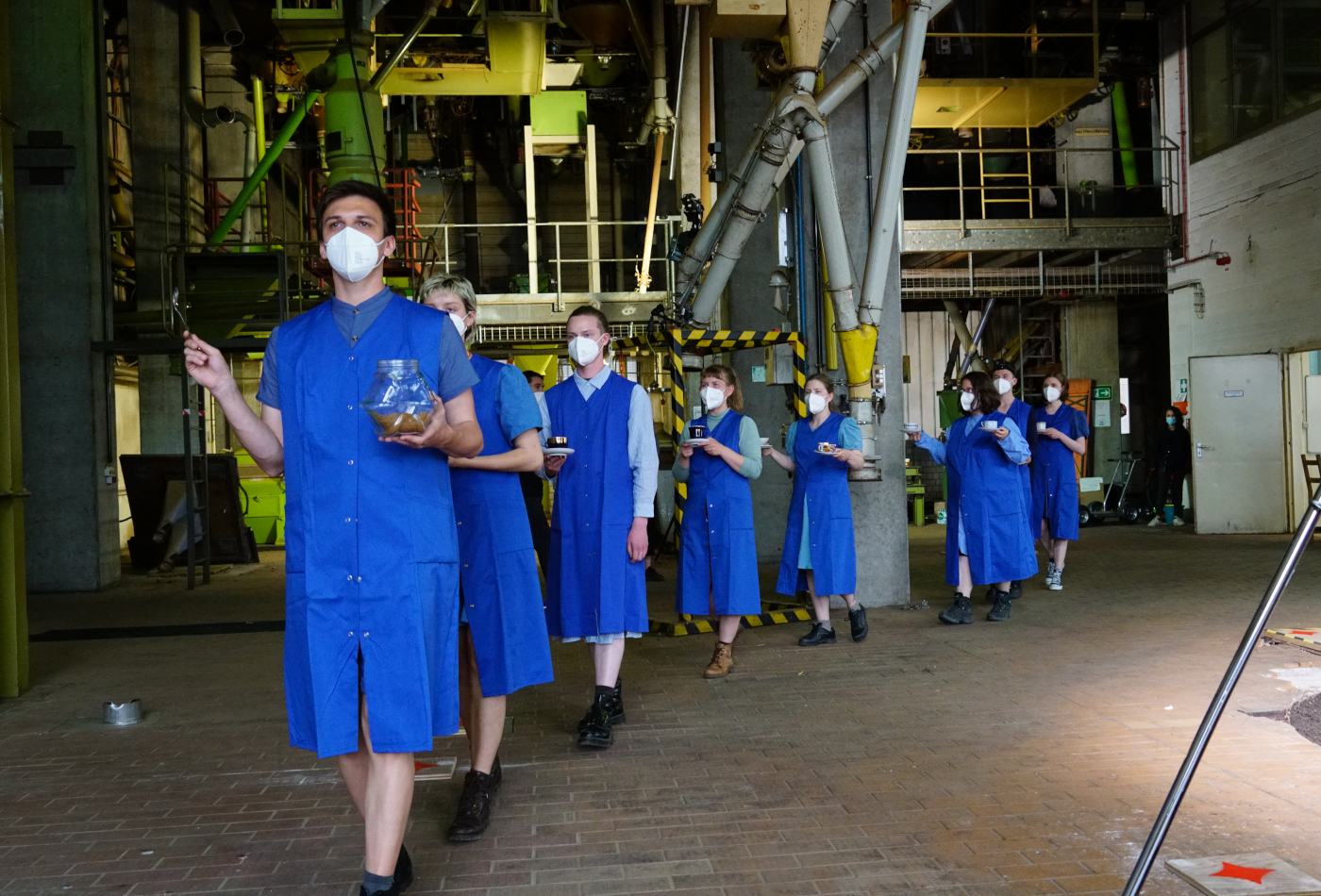 Personen mit blauen Kitteln laufen durch eine Produktionshalle.