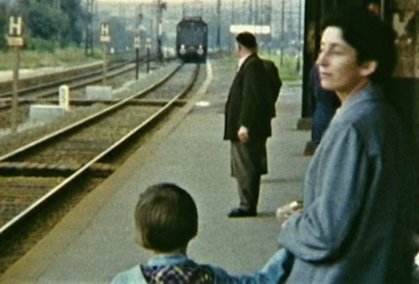 Eine Frau steht mit einem Jungen auf dem Gleis. Ein Zug fährt ein.