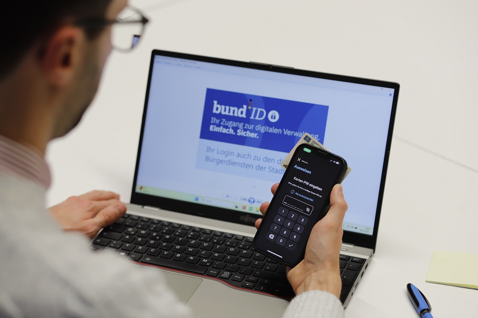 Mann vor Laptop. Internetseite der Bund ID geöffnet, hier ist Login zu den digitalen Bürgerdiensten der Stadtverwaltung möglich. In der Hand hält er einen Personalausweis und ein Smartphone, auf dem die AusweisApp geöffnet ist. Die App fordert ihn auf, eine PIN einzugeben.ben