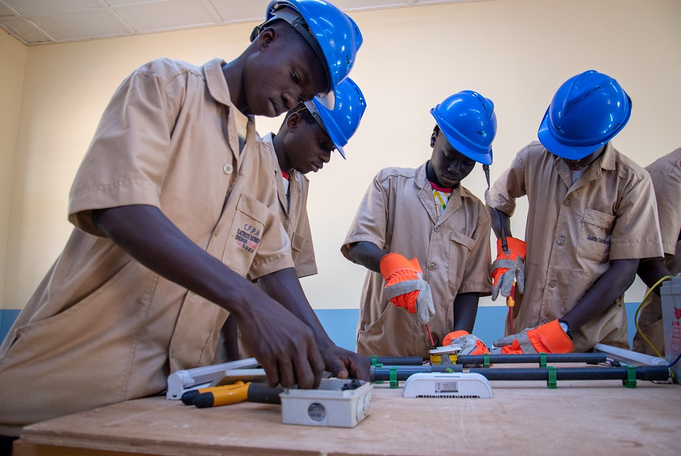 Vier Jugendliche in Arbeitskleidung und mit Schutzhelmen montieren technische Bauteile an einem Tisch.